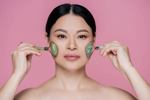 Asian woman with naked shoulders massaging cheeks with jade rollers isolated on pink — Stock Photo