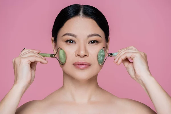 Femme asiatique aux épaules nues massant les joues avec des rouleaux de jade isolés sur rose — Photo de stock