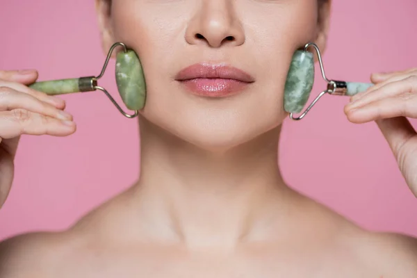 Vista recortada de mujeres masajeando mejillas con rodillos de jade aislados en rosa - foto de stock