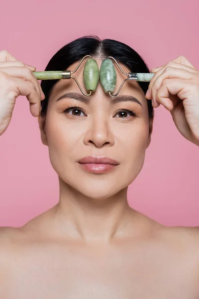 Asian woman with naked shoulders massaging forehead with jade rollers isolated on pink — Stock Photo