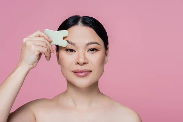 Asian woman with naked shoulders scraping face with jade stone isolated on pink — Stock Photo