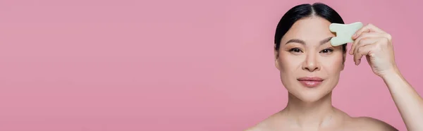 Азиатка с обнаженным плечом массирует лицо нефритовым камнем, изолированным на розовом, баннер — стоковое фото