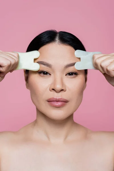 Asiatische Frau mit nackten Schultern, die Jadesteine in der Nähe von Augenbrauen hält, isoliert auf rosa — Stockfoto