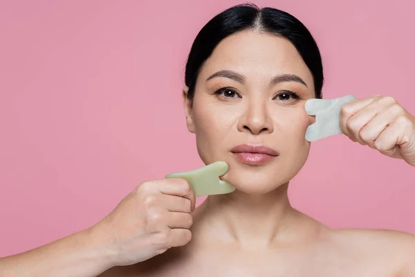 Asiatische Frau kratzt Gesicht mit Jadesteinen und schaut Kamera isoliert auf rosa — Stockfoto