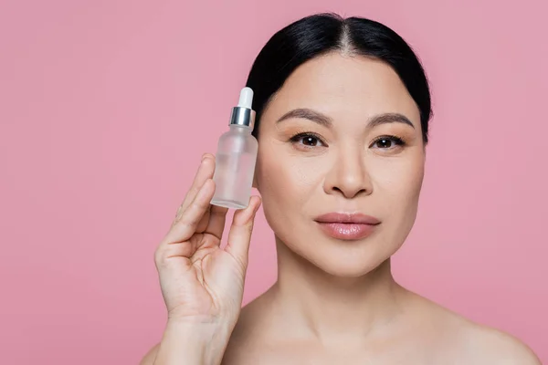 Femme asiatique tenant bouteille avec sérum cosmétique isolé sur rose — Photo de stock