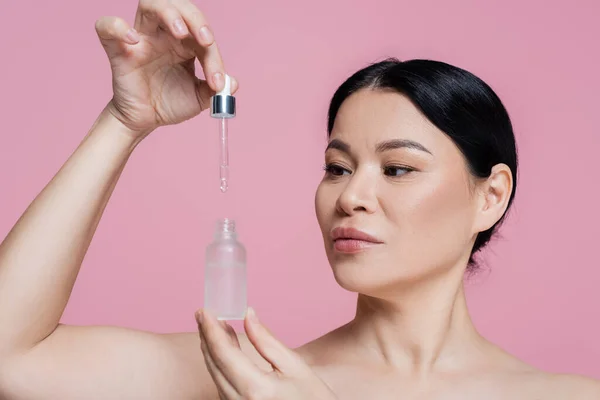 Asian woman with naked shoulders holding dropper and serum isolated on pink — Stock Photo