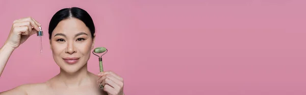 Asian woman with naked shoulders holding jade roller and serum isolated on pink, banner — Stock Photo