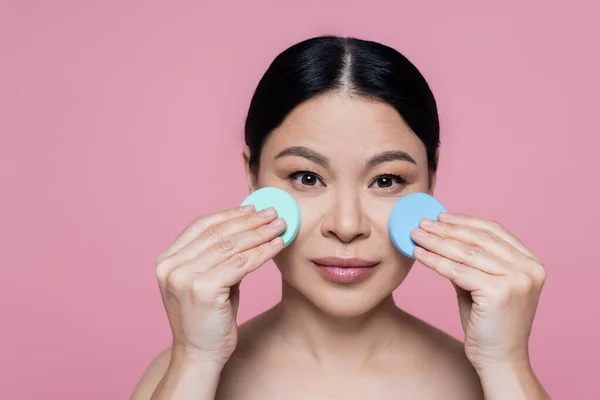 Asian woman with naked shoulders cleaning face with sponges isolated on pink — Stock Photo