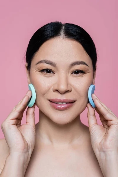Smiling asian woman cleaning face with sponges isolated on pink — Stock Photo