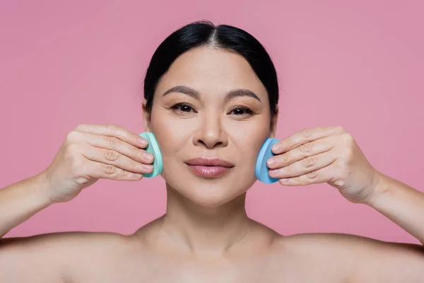 Asian woman with naked shoulders cleaning face with sponges isolated on pink — Stock Photo