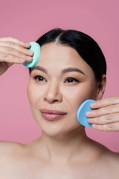 Asiatische Frau mit nackten Schultern putzt Gesicht mit Schwämmen isoliert auf rosa — Stockfoto