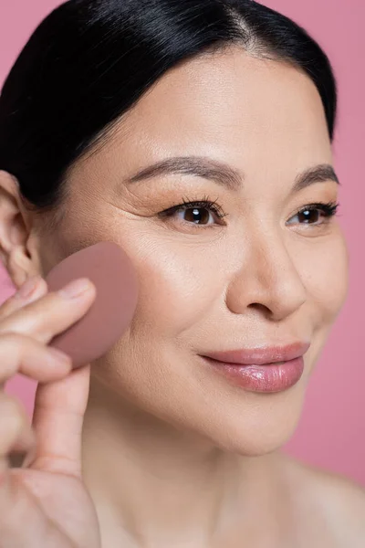 Close up view of asian woman applying face foundation with blurred beauty blender isolated on pink — Stock Photo