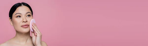 Азиатка с обнаженными плечами очищает лицо губкой, изолированной на розовом, баннер — стоковое фото