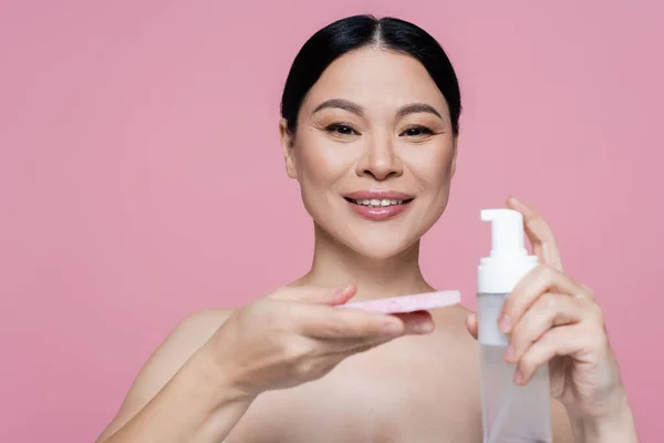 Femme asiatique positive avec des épaules nues tenant éponge floue et mousse nettoyante isolée sur rose — Photo de stock