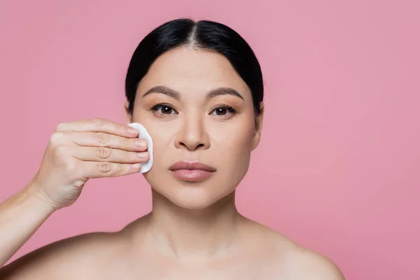 Mulher asiática com ombros nus limpeza rosto com almofada de algodão isolado em rosa — Fotografia de Stock