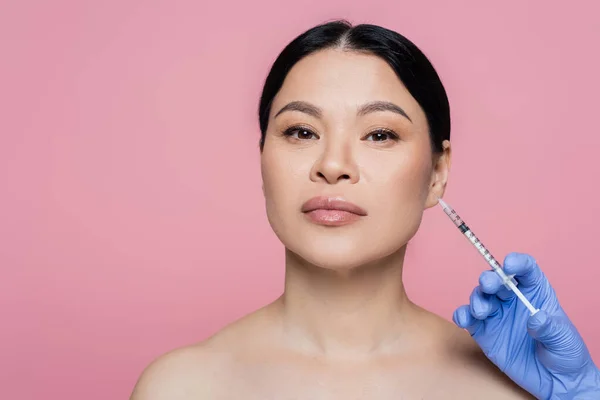 Cosmetologist in latex glove holding syringe near face of asian woman isolated on pink — Stock Photo