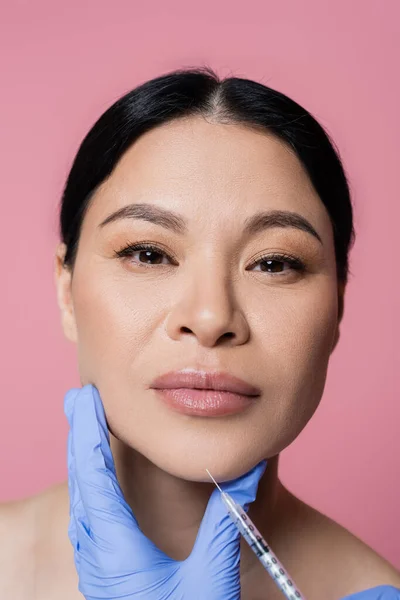 Cosmetologue tenant la seringue près du menton de la femme asiatique regardant la caméra isolée sur rose — Photo de stock