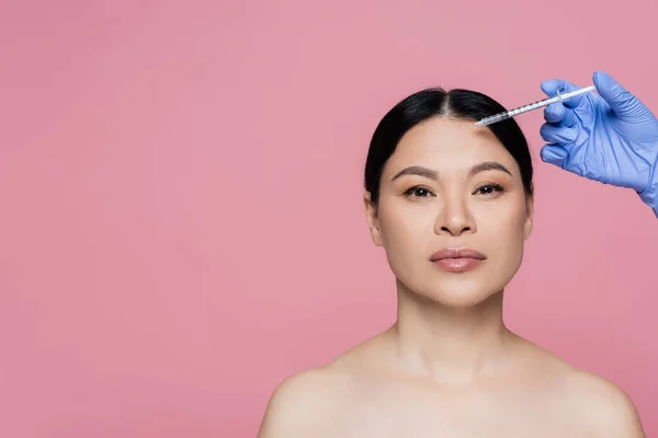 Cosmetólogo en guante de látex sosteniendo jeringa cerca de la frente de una mujer asiática aislada en rosa - foto de stock