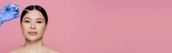 Estetista tenuta siringa vicino al viso di donna asiatica con spalle nude isolate su rosa, banner — Foto stock
