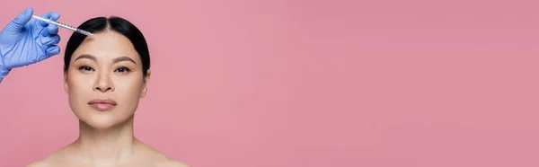Beautician holding syringe near face of asian woman with naked shoulders isolated on pink, banner — Stock Photo