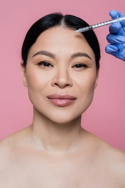 Mujer asiática con hombros desnudos mirando a la cámara cerca del cosmetólogo sosteniendo la jeringa aislada en rosa - foto de stock