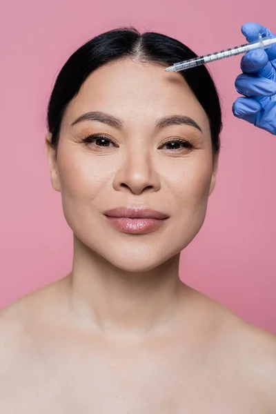 Asian woman with naked shoulders looking at camera near cosmetologist holding syringe isolated on pink — Stock Photo