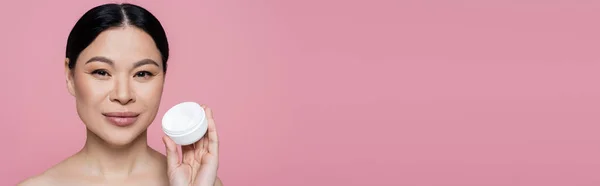 Donna asiatica con spalle nude che tiene il contenitore con crema cosmetica isolata su rosa, banner — Foto stock