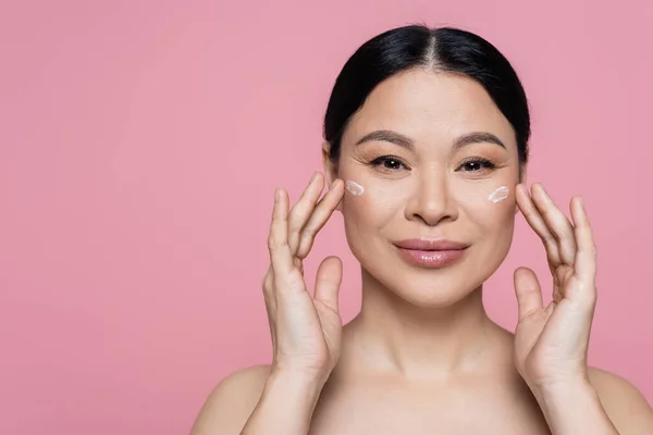Cheerful asian woman applying face cream on cheeks isolated on pink — Stock Photo