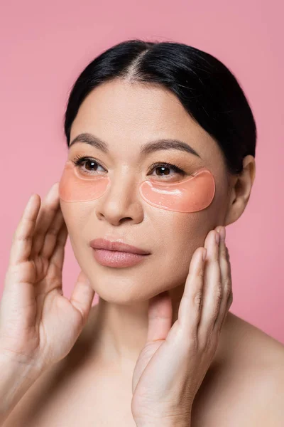 Mujer asiática con parches de hidrogel en los ojos tocando la cara aislada en rosa - foto de stock