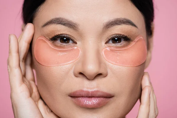 Vista de cerca de la mujer asiática con parches en los ojos tocando la cara aislado en rosa - foto de stock