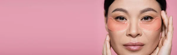 Femme asiatique avec des patchs hydrogel pour les yeux regardant la caméra isolée sur rose, bannière — Photo de stock