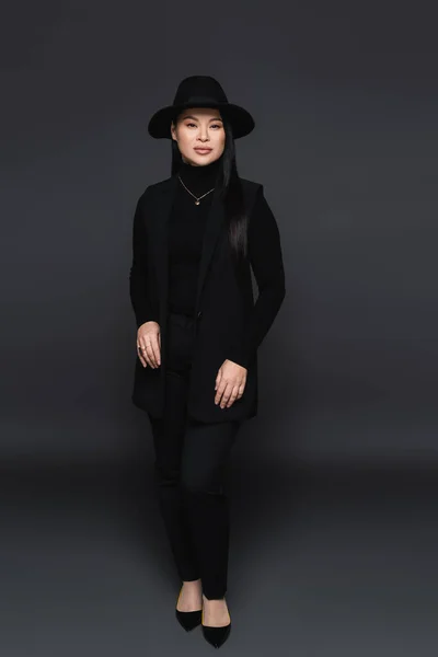 Полная длина стильной азиатской женщины, смотрящей на камеру на темно-сером фоне — стоковое фото