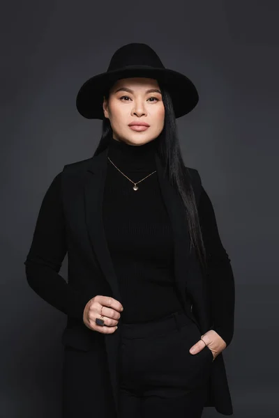 Élégante femme asiatique en chapeau fedora tenant la main dans la poche de pantalon isolé sur gris foncé — Photo de stock