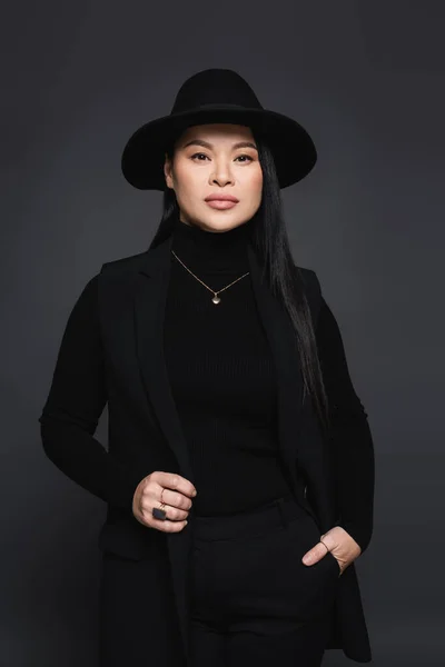 Elegant asian woman in fedora hat holding hand in pocket of pants isolated on dark grey — Stock Photo