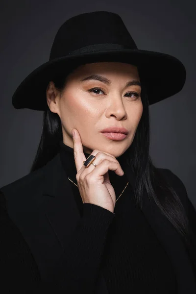 Retrato de mujer asiática de moda en sombrero fedora mirando a la cámara aislada en gris oscuro - foto de stock