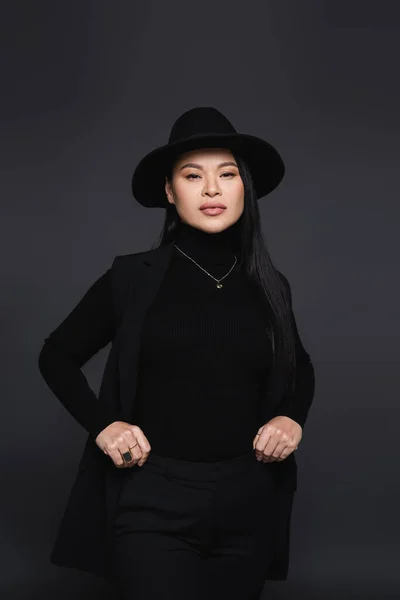Elegante mujer asiática en sombrero fedora y cuello alto mirando a la cámara aislada en gris oscuro — Stock Photo
