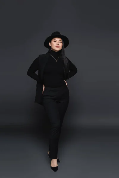 Full length of stylish asian woman in fedora hat holding hands on hips on dark grey background — Stock Photo