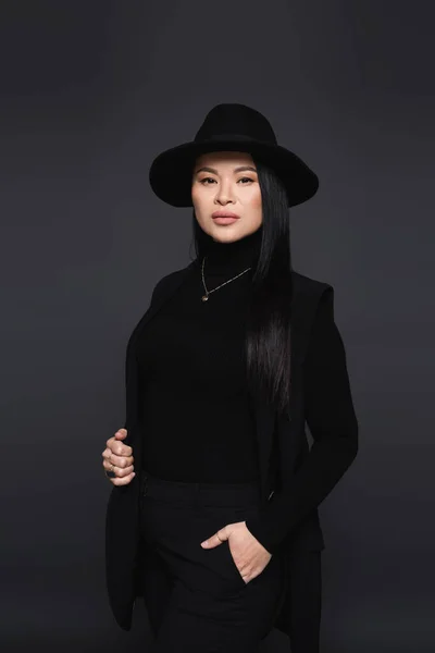 Stylish asian woman in fedora hat posing and looking at camera isolated on dark grey — Stock Photo