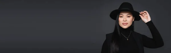 Modelo asiático tocando sombrero fedora y mirando a la cámara aislada en gris oscuro, pancarta - foto de stock