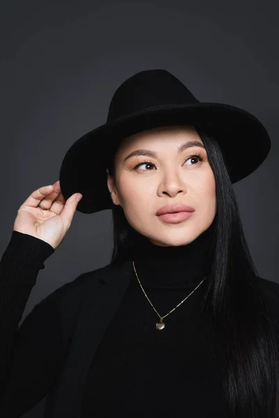 Retrato de mujer asiática elegante ajustando sombrero fedora aislado en gris oscuro - foto de stock