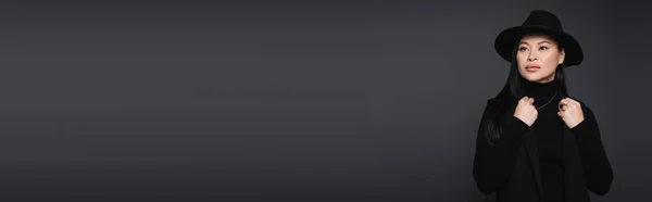 Азиатская модель в шляпе Fedora и водолазке позирует изолированно на темно-сером, баннер — стоковое фото