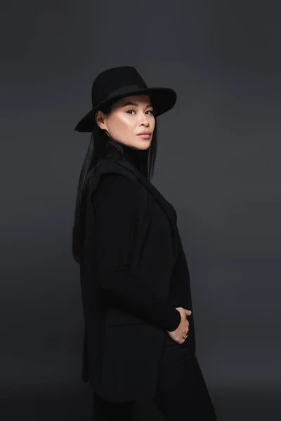 Fashionable asian woman in fedora hat looking at camera isolated on dark grey — Stock Photo