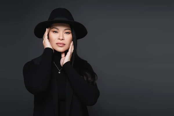 Stylish asian woman in fedora hat touching face isolated on dark grey — Stock Photo