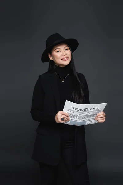 Cheerful asian woman in fedora hat and jacket holding newspaper isolated on dark grey — Stock Photo