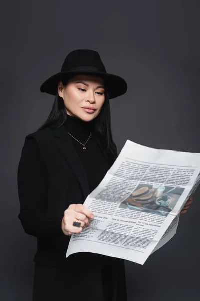 Asian woman in fedora hat and blazer reading newspaper isolated on dark grey — Stock Photo