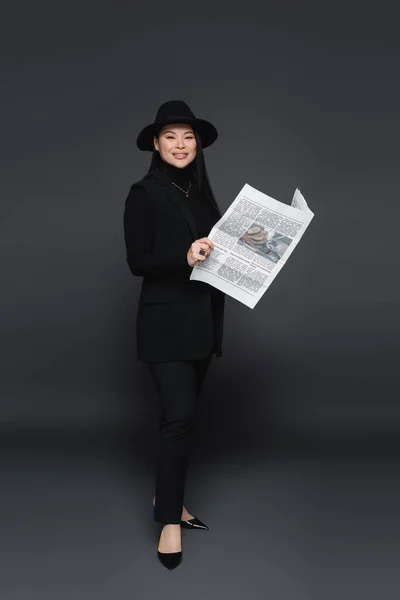 Полная длина улыбающейся азиатской модели в шляпе-шляпе, держащей газету на темно-сером фоне — стоковое фото