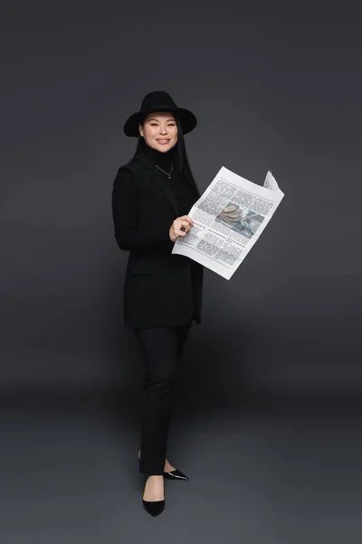 Full length of smiling asian model in fedora hat holding newspaper on dark grey background — Stock Photo