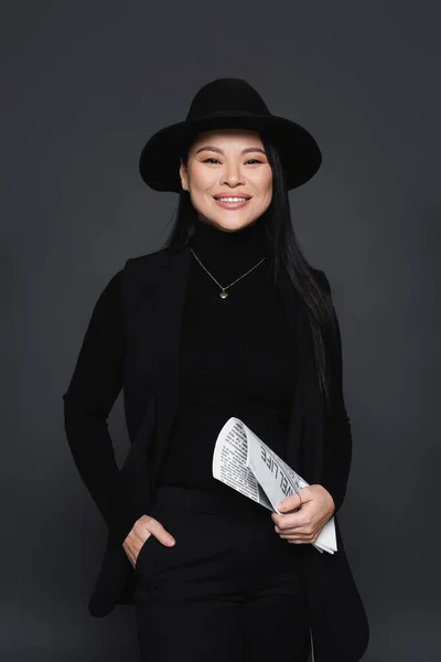 Sorrindo mulher asiática em blazer e chapéu fedora segurando jornal isolado no cinza escuro — Fotografia de Stock