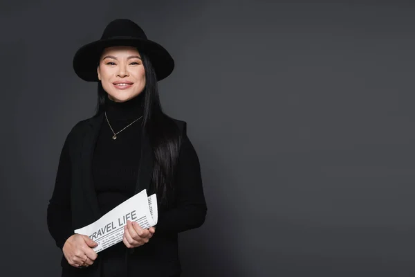 Mujer con estilo positivo en sombrero fedora sosteniendo periódico aislado en gris oscuro - foto de stock