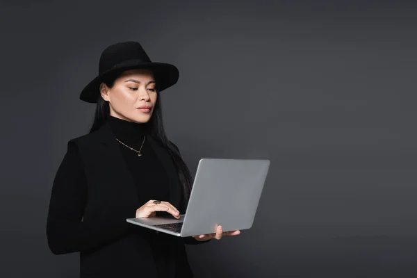 Stylish asian woman in fedora hat using laptop isolated on dark grey — Stock Photo